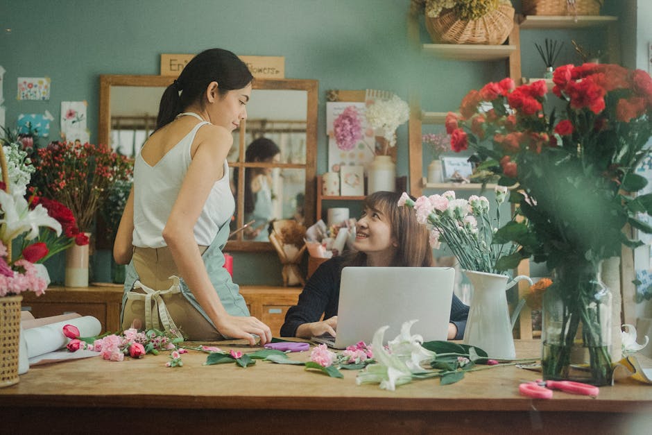 A florist and a customer in a flower shop engaged in a conversation, with the florist standing and leaning slightly over the wooden table, smiling at the customer who is seated behind a laptop. The ta