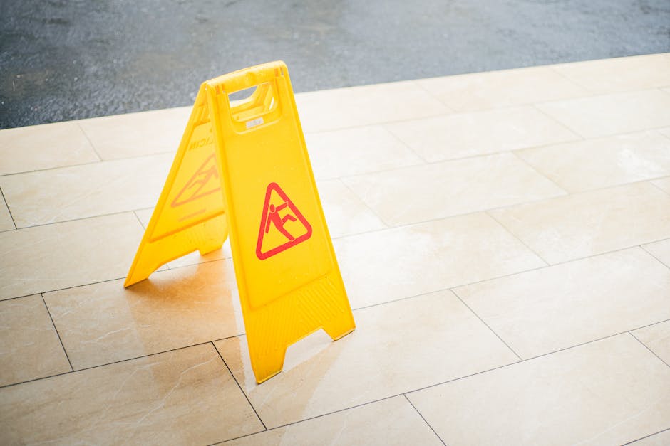 A bright yellow, portable wet floor warning sign with red pictograms and text, placed on a light beige tiled floor near a dark surface, serving as a caution for potential slipping hazards. The sign is
