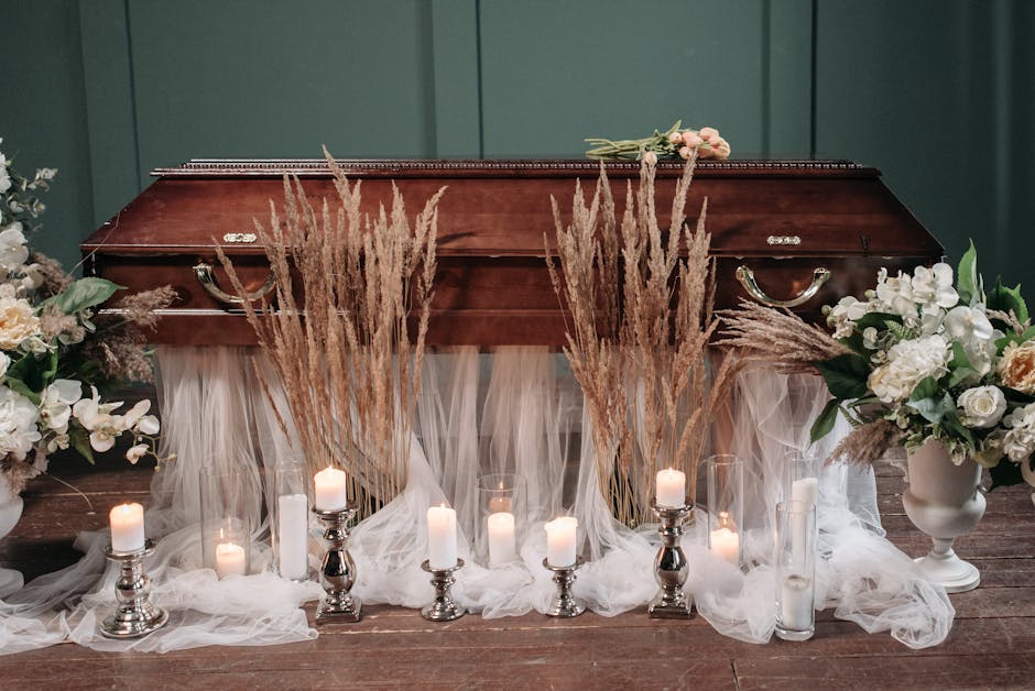 A funeral floral arrangement displayed in front of a closed wooden casket, featuring tall, beige pampas grass and white flowers in elegant white vases. The flowers include roses and other delicate blo