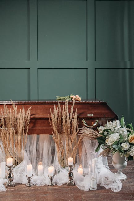 A floral arrangement featuring white roses and green foliage in a white ceramic vase, placed on a wooden surface with soft, neutral-colored fabric draped around it. The setup includes a row of small w