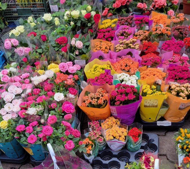 A colorful display of fresh flowers at a flower shop, including pink and white roses, various carnations, and small-flowered arrangements wrapped in bright yellow, purple, orange, and pink paper. The