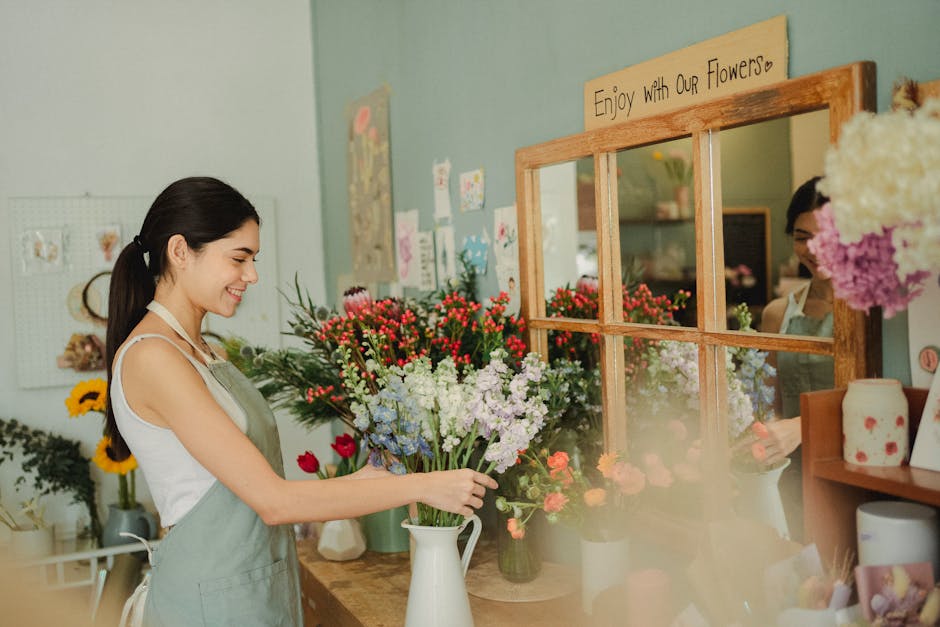 A young woman with long dark hair tied back, smiling warmly as she arranges a vibrant bouquet of fresh flowers in a white ceramic pitcher inside a flower shop. The bouquet includes purple, red, and wh