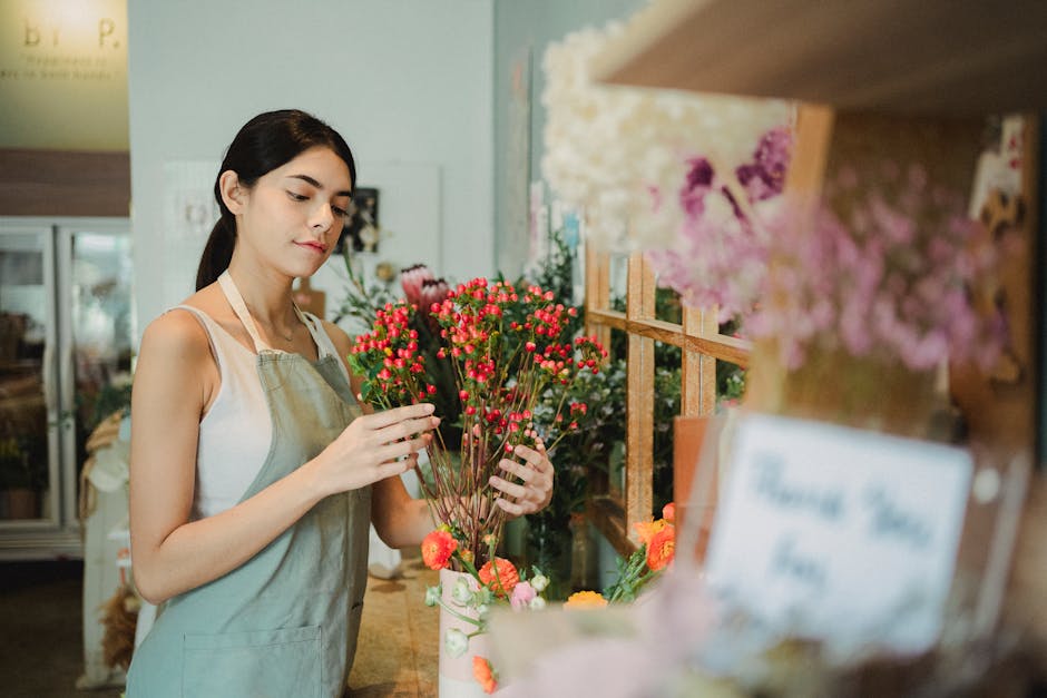 A young woman with dark hair tied back, dressed in a light-colored sleeveless top and a beige apron, stands inside a flower shop, carefully arranging a bouquet of small, vibrant pink and red waxflower
