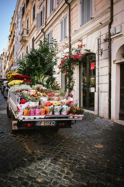 A flatbed truck parked on a cobblestone street in front of a cream-colored building with large windows and a glass door, filled with an array of colorful fresh flowers arranged in bouquets, potted pla