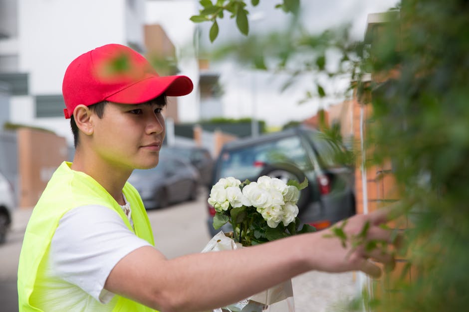 A delivery person wearing a red cap and a high-visibility yellow vest is placing a floral bouquet of white roses and hydrangeas wrapped in light paper onto a doorstep. The arrangement is fresh, with v
