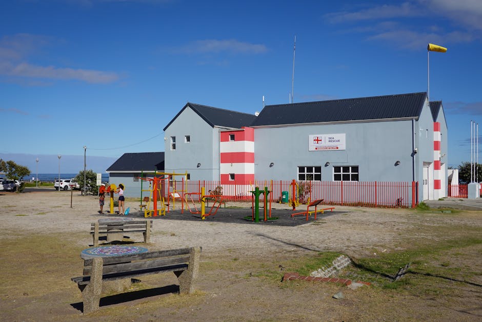 A seaside building painted in light blue with red and white striped accents, housing a Sea Rescue center, situated behind an outdoor children's playground with colorful equipment and two children play