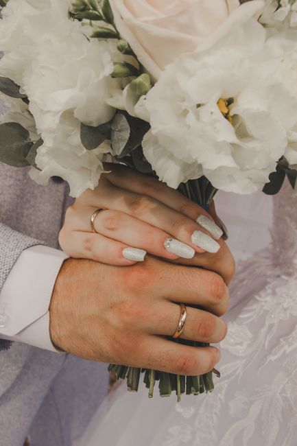 Close-up of a bride and groom holding a bouquet of fresh white flowers, including eustoma and lisianthus, wrapped in dark green foliage. The bride's hand, with white glittery nail polish, rests over t